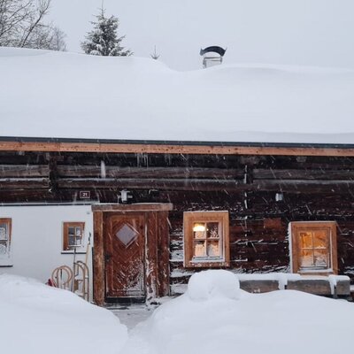 Die Alm im Winter, mit dickem Schnee auf dem Dach und dem umliegenden Gelände, beleuchteten Fenstern und bereitstehenden Schlitten am Eingang.