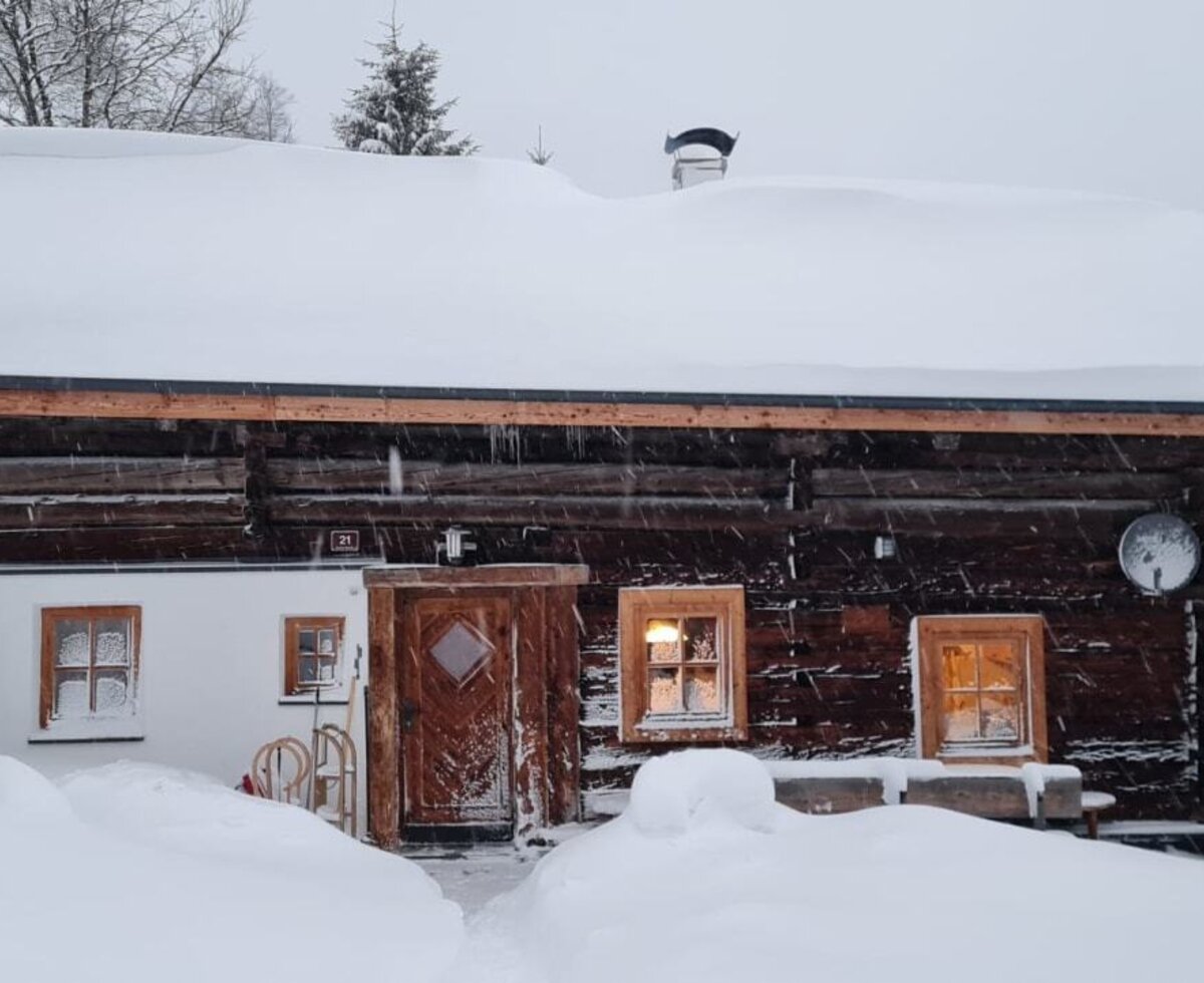 Die Alm im Winter, mit dickem Schnee auf dem Dach und dem umliegenden Gelände, beleuchteten Fenstern und bereitstehenden Schlitten am Eingang.
