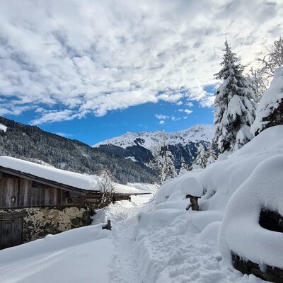 Die verschneite Alm mit Stein- und Holzfassade, umgeben von tiefem Schnee und einem Winterwanderweg in den Bergen.