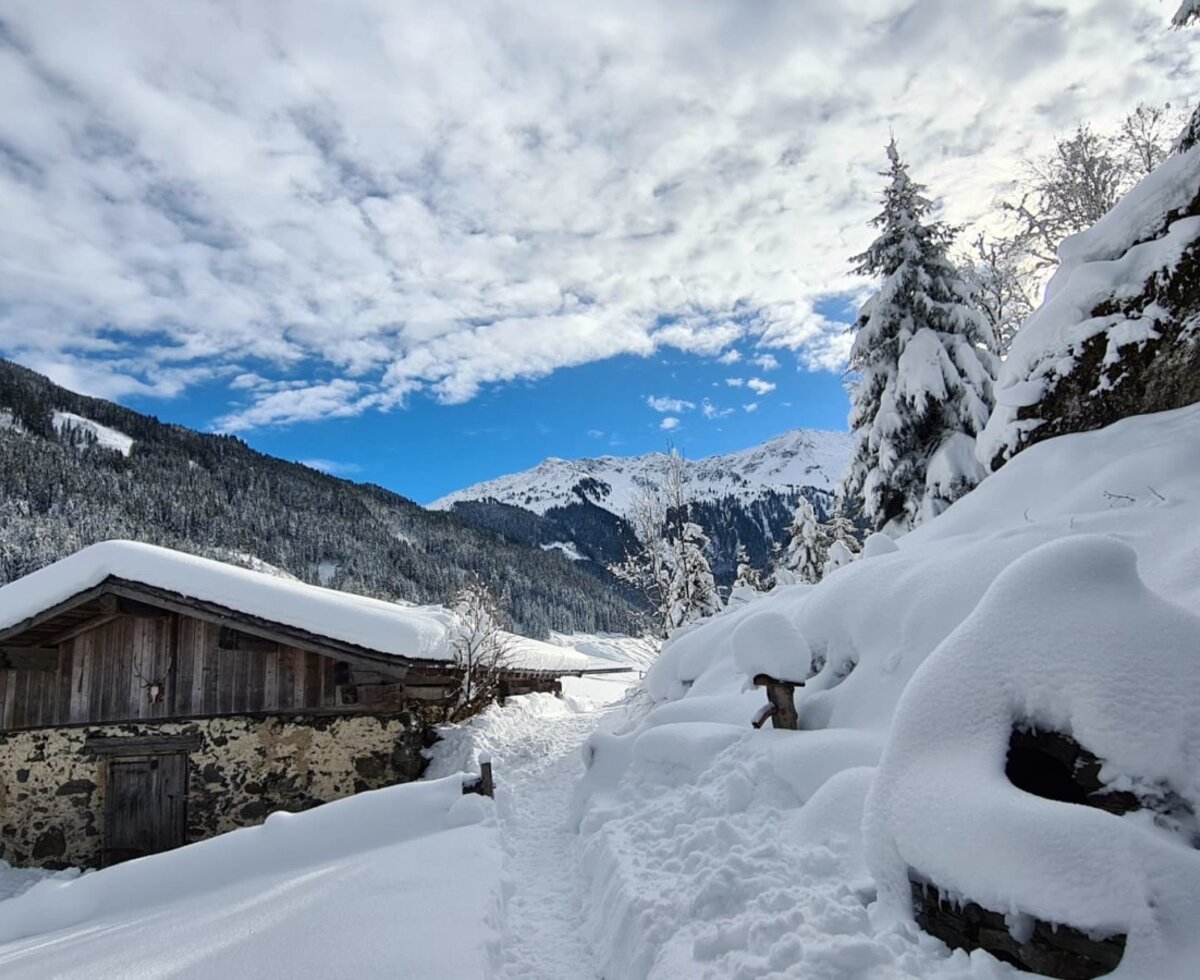 Die verschneite Alm mit Stein- und Holzfassade, umgeben von tiefem Schnee und einem Winterwanderweg in den Bergen.