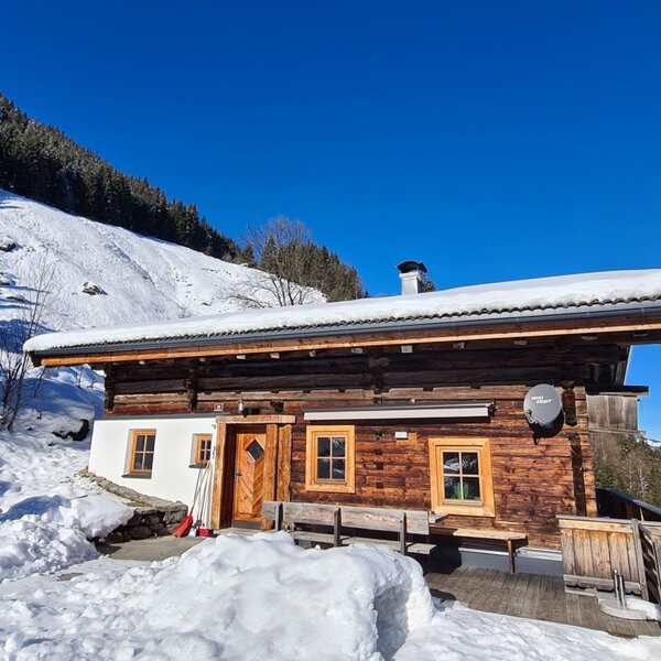 Die Alm mit Holzfassade und schneebedecktem Dach, umgeben von verschneiten Bergen unter blauem Himmel, mit einem Außenofen.
