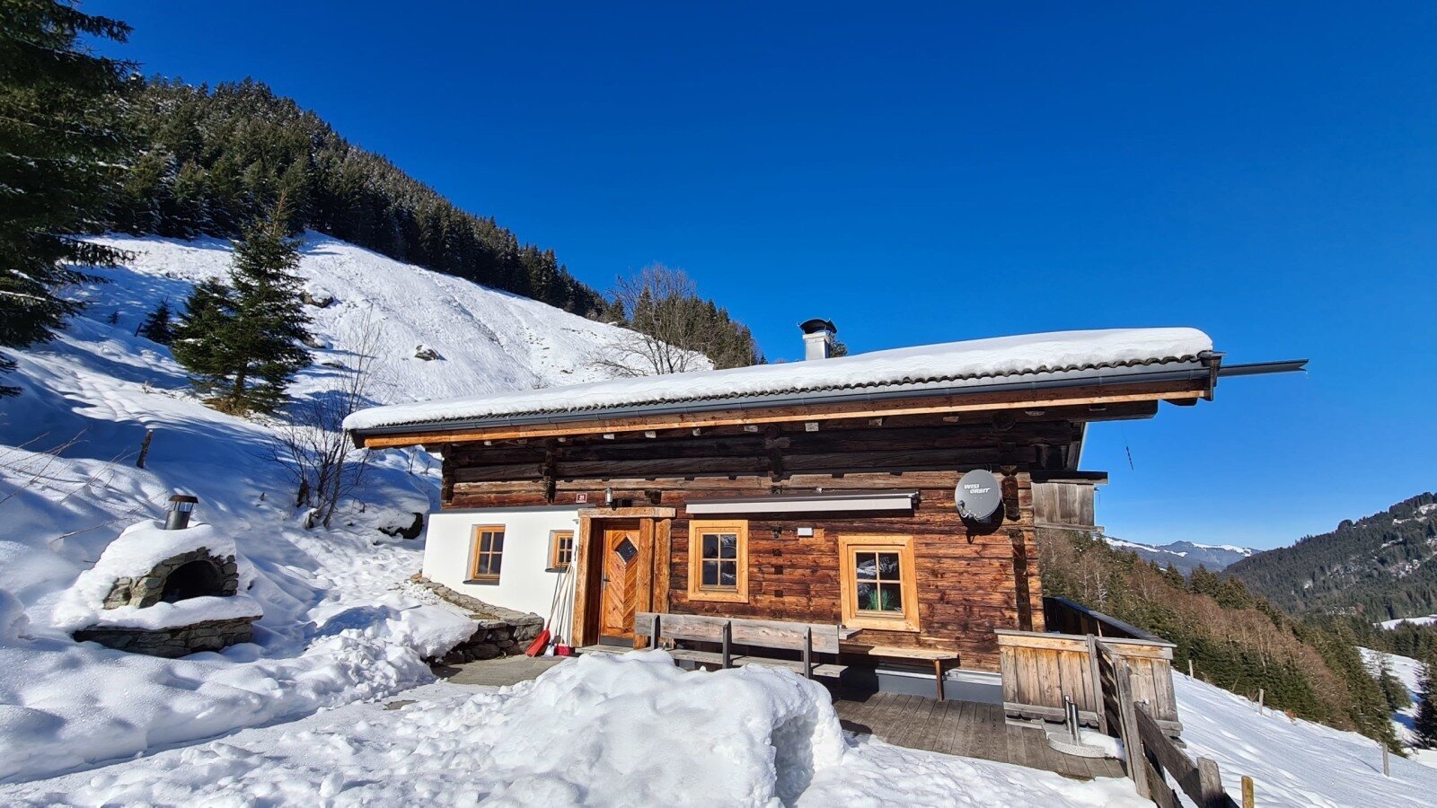 Die Alm mit Holzfassade und schneebedecktem Dach, umgeben von verschneiten Bergen unter blauem Himmel, mit einem Außenofen.