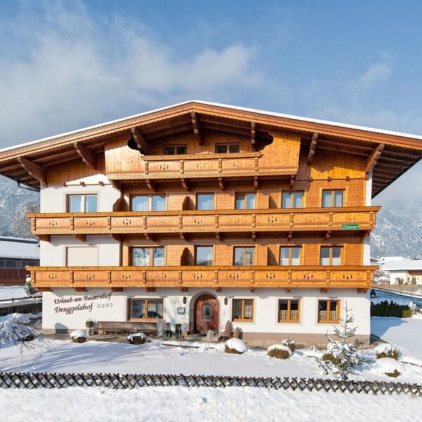 Außenansicht des Bauernhofs mit traditionellen Holzbalkonen, umgeben von Schnee und den Alpen.