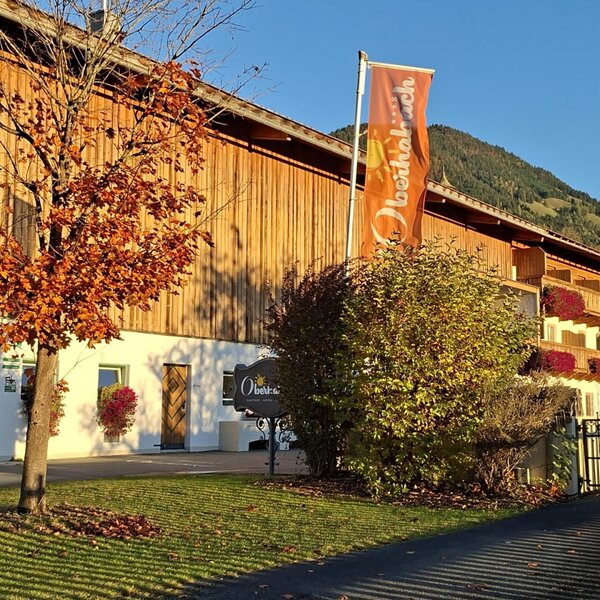 Außenansicht des Bauernhofs mit Holzfassade, Balkonen mit Blumen und einem Baum mit Herbstlaub.