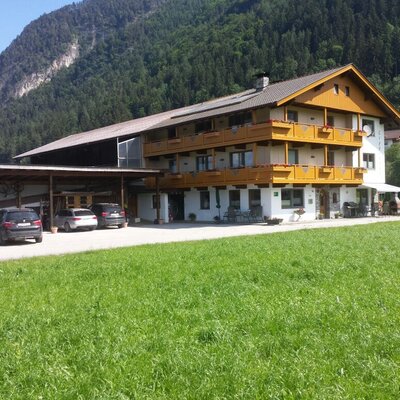 Der Bauernhof mit Holzverkleidung und Balkonen, angrenzendem Unterstand für Fahrzeuge und umgeben von grünen Wiesen und Bergen.