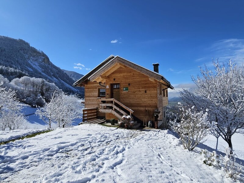 Das hölzerne Ferienhaus auf dem Hof, umgeben von Schnee und Bergen unter einem klaren blauen Himmel.