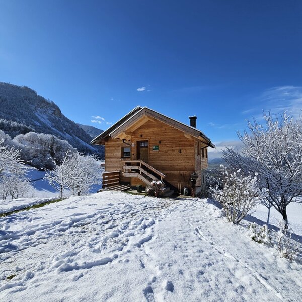 Das hölzerne Ferienhaus auf dem Hof, umgeben von Schnee und Bergen unter einem klaren blauen Himmel.