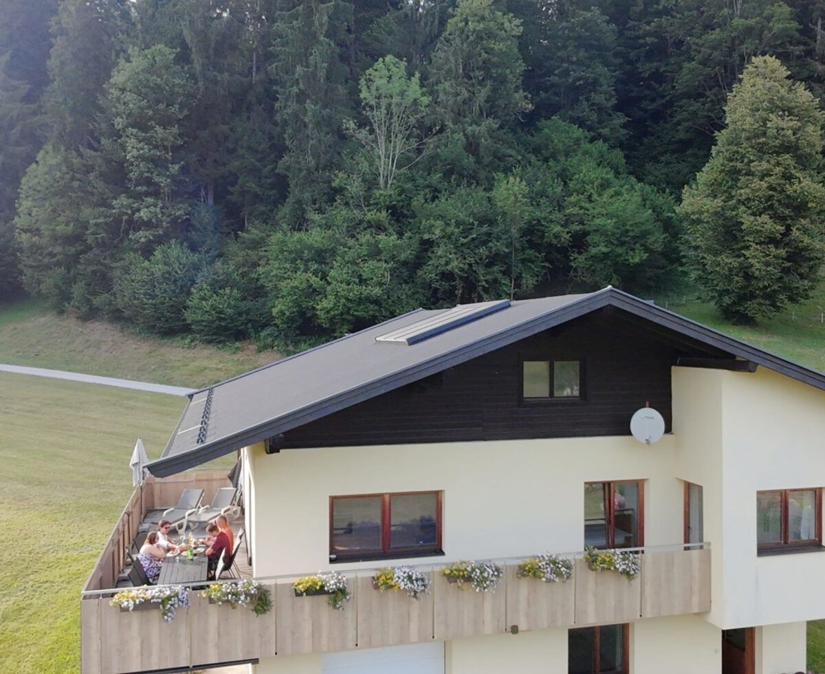Das einladende Bauernhaus mit einem großen, blumengeschmückten Balkon, umgeben von grünen Wiesen und Wäldern.