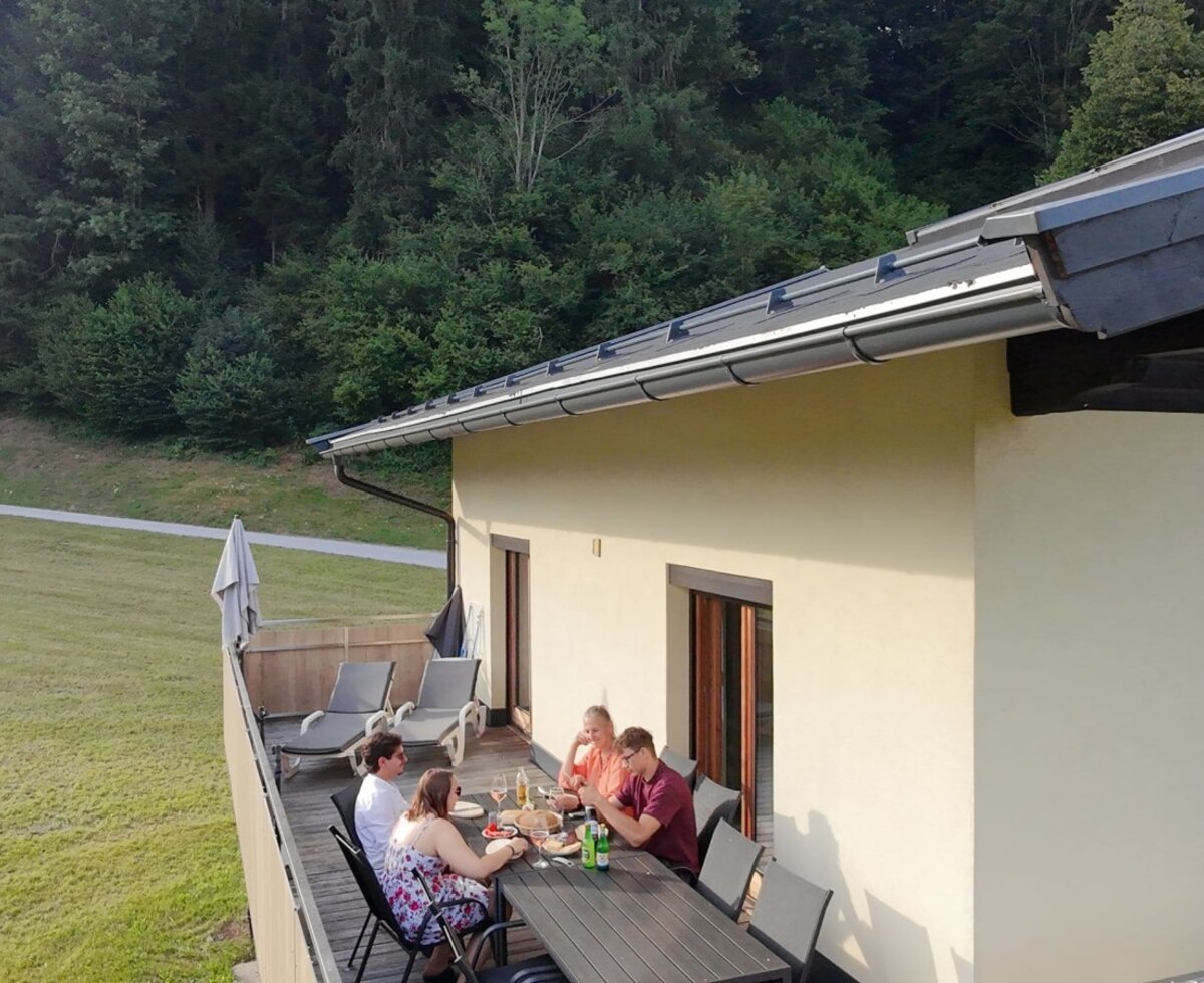 Die sonnige Terrasse des Bauernhofs mit einem großen Esstisch und Sonnenliegen vor einer idyllischen Waldkulisse.