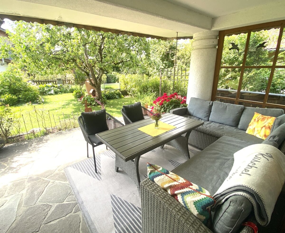 Die einladende, überdachte Terrasse des Bauernhofs mit einer gemütlichen Sitzlandschaft, einem Esstisch und Blick in den grünen Garten.
