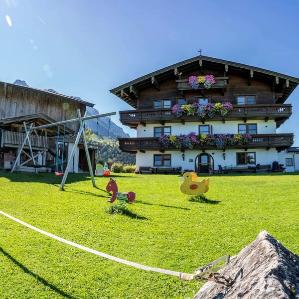 Außenansicht des Bauernhofs mit blumengeschmückten Balkonen, einem großen Rasen mit Kinderspielplatz und einer Slackline.