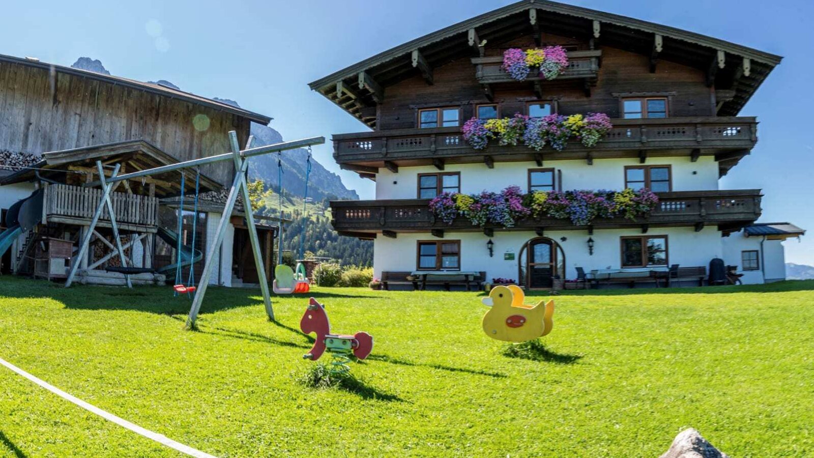 Außenansicht des Bauernhofs mit blumengeschmückten Balkonen, einem großen Rasen mit Kinderspielplatz und einer Slackline.