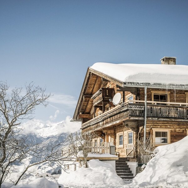 Der Bauernhof im Winter, ein Holzgebäude mit Balkonen, Satellitenanlage und verschneitem Dach, umgeben von schneebedeckten Bergen.