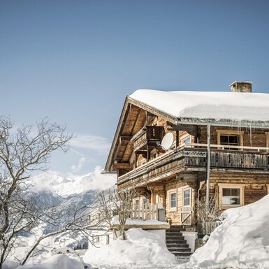 Der Bauernhof im Winter, ein Holzgebäude mit Balkonen, Satellitenanlage und verschneitem Dach, umgeben von schneebedeckten Bergen.
