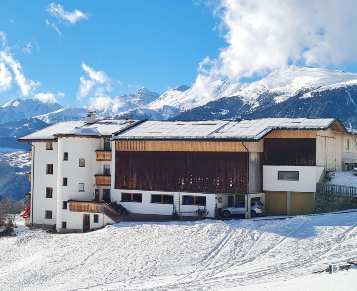 Außenansicht des Bauernhofs mit weißer Fassade, Holzbalkonen und schneebedecktem Dach, umgeben von Winterlandschaft und Bergen.