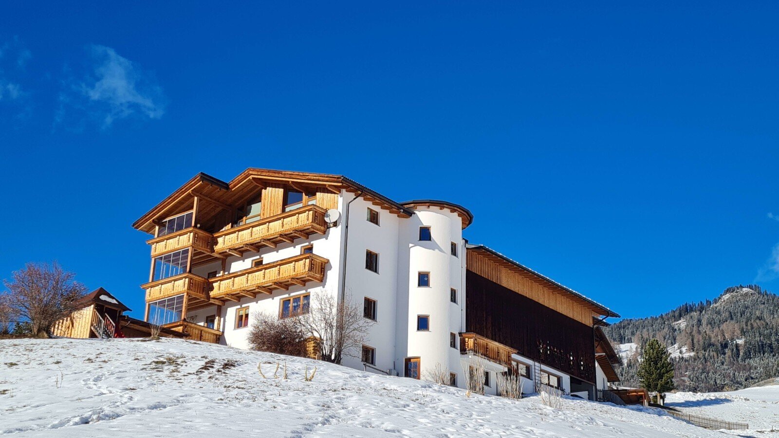 Der Bauernhof im Winter, mit weißer Fassade, Holzverkleidung und Balkonen, auf einem schneebedeckten Hügel.
