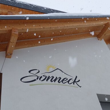 Der Name und das Logo des Bauernhofs Sonneck an der Außenfassade, mit sichtbaren Holzelementen unter dem Dach und fallendem Schnee.