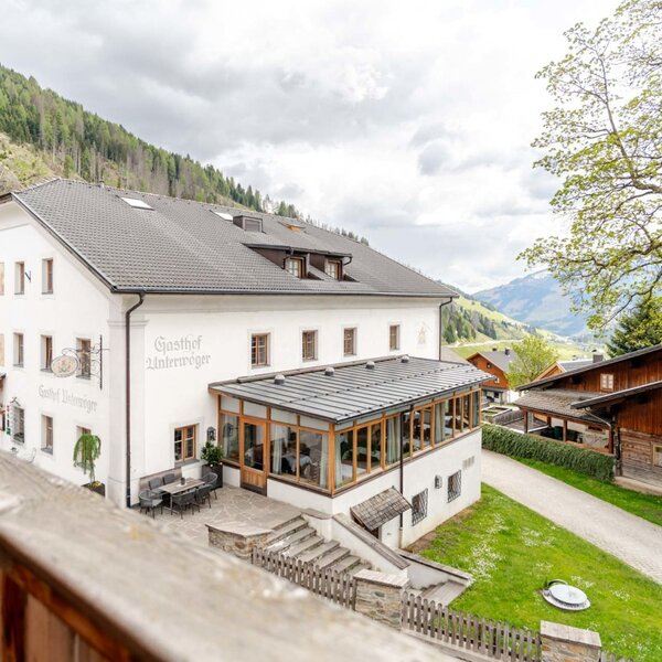 Außenansicht des Bauernhofs mit einem weißen Hauptgebäude, einem verglasten Essbereich und einer Außenterrasse, umgeben von Bergen.