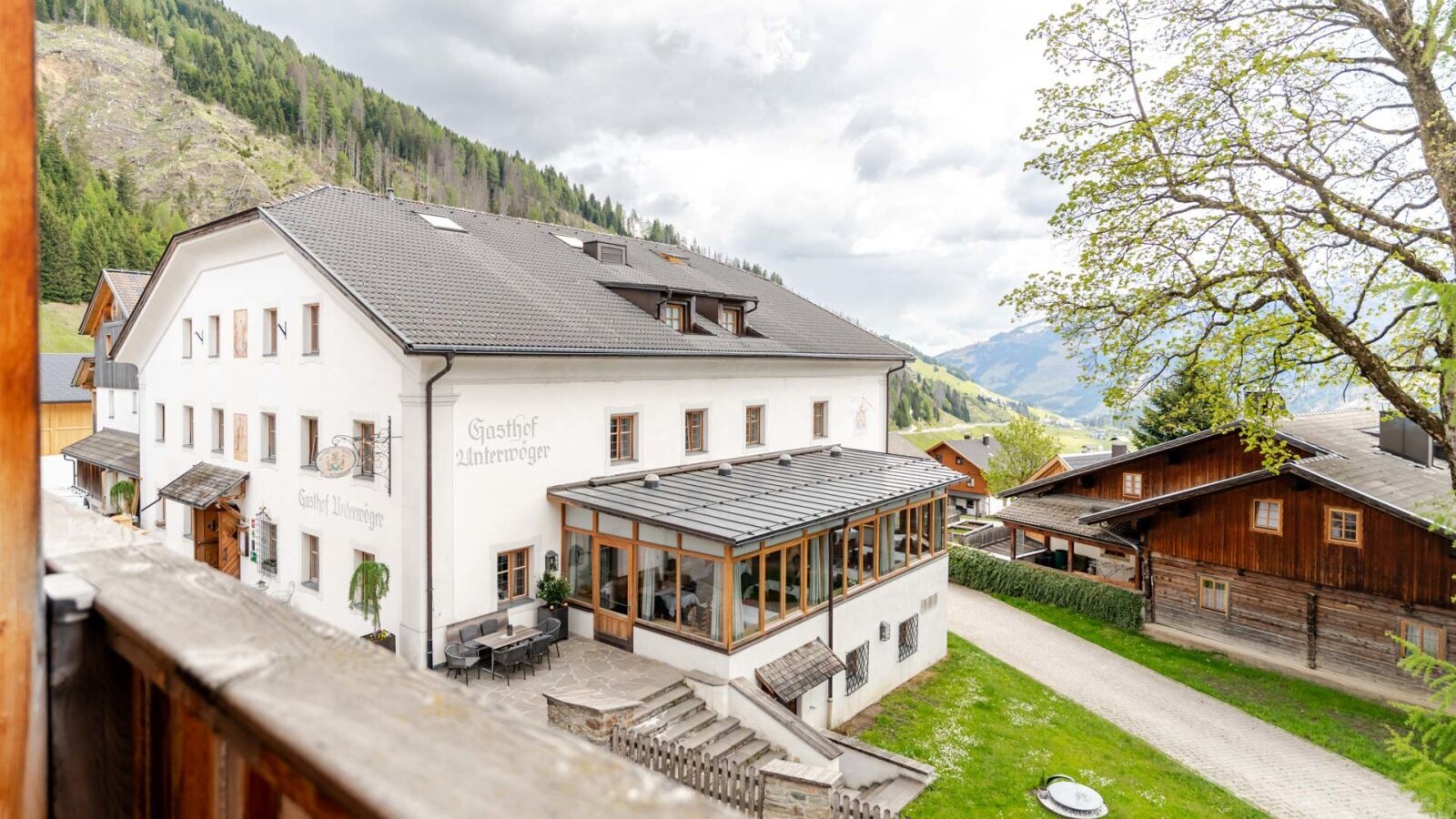 Außenansicht des Bauernhofs mit einem weißen Hauptgebäude, einem verglasten Essbereich und einer Außenterrasse, umgeben von Bergen.