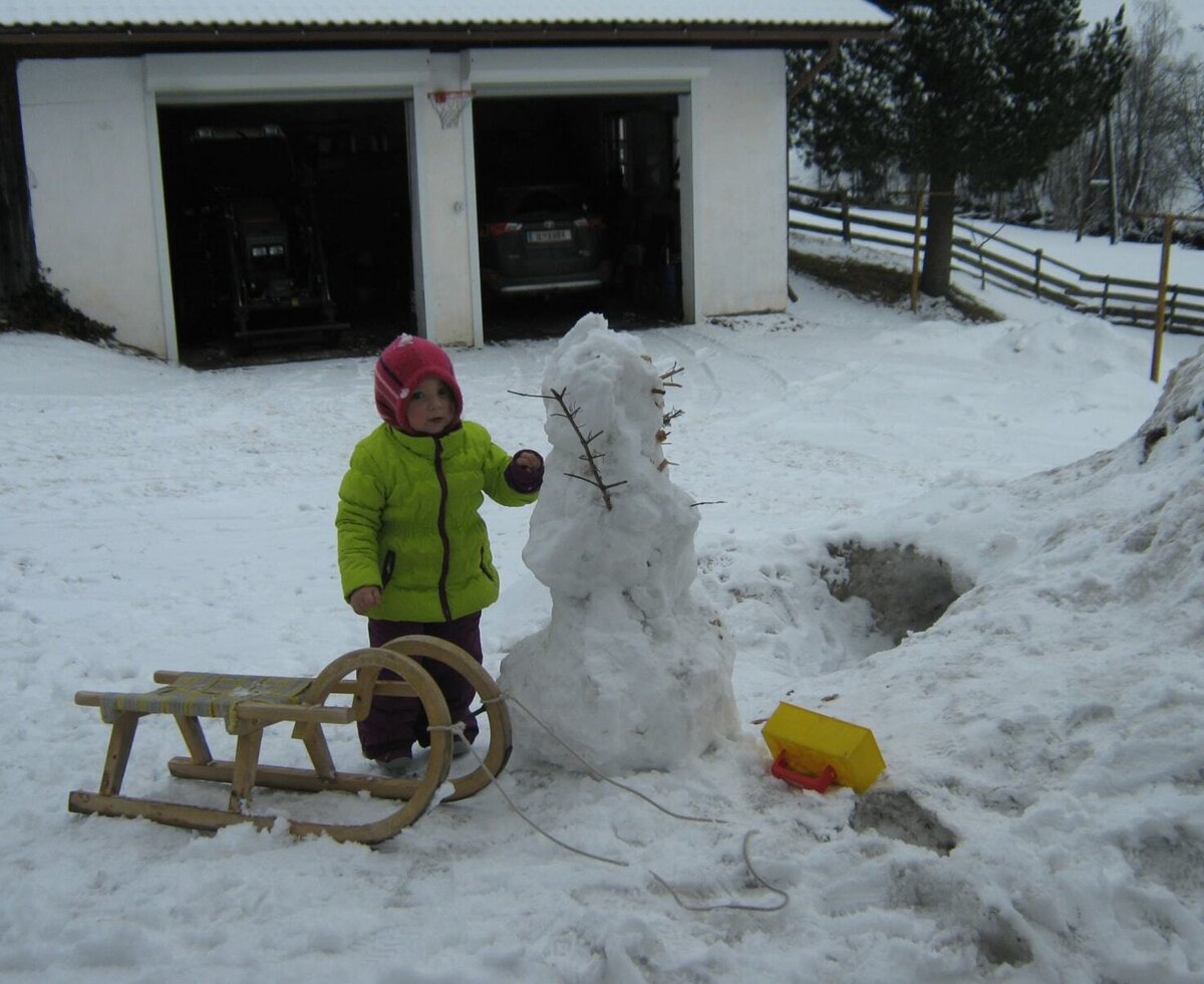 Ein Kind spielt mit einem Schneemann und einem Schlitten im Schnee auf dem Bauernhof.