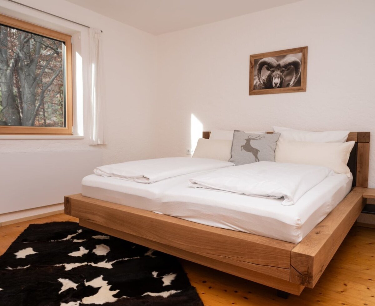 Schlafzimmer im Apartment mit einem Doppelbett, Holzboden, einem schwarz-weißen Teppich und einem Fenster mit Blick ins Grüne.