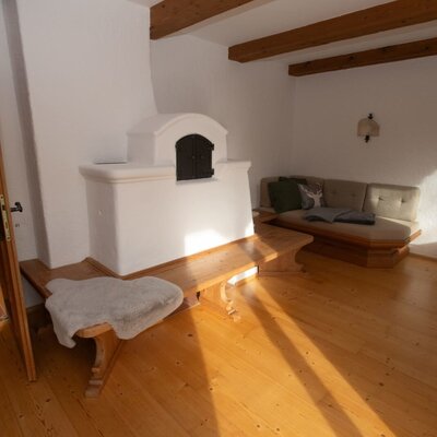 Wohnbereich des Apartments mit traditionellem Kachelofen, Holzbank, Sofa und Holzbalkendecke.