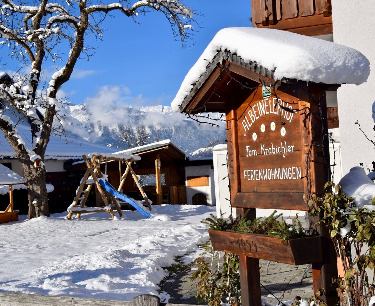 Das Willkommensschild des Bauernhofs "AlbeinelERhof" im Schnee, mit Kinderspielplatz und Bergblick auf die Alpen.