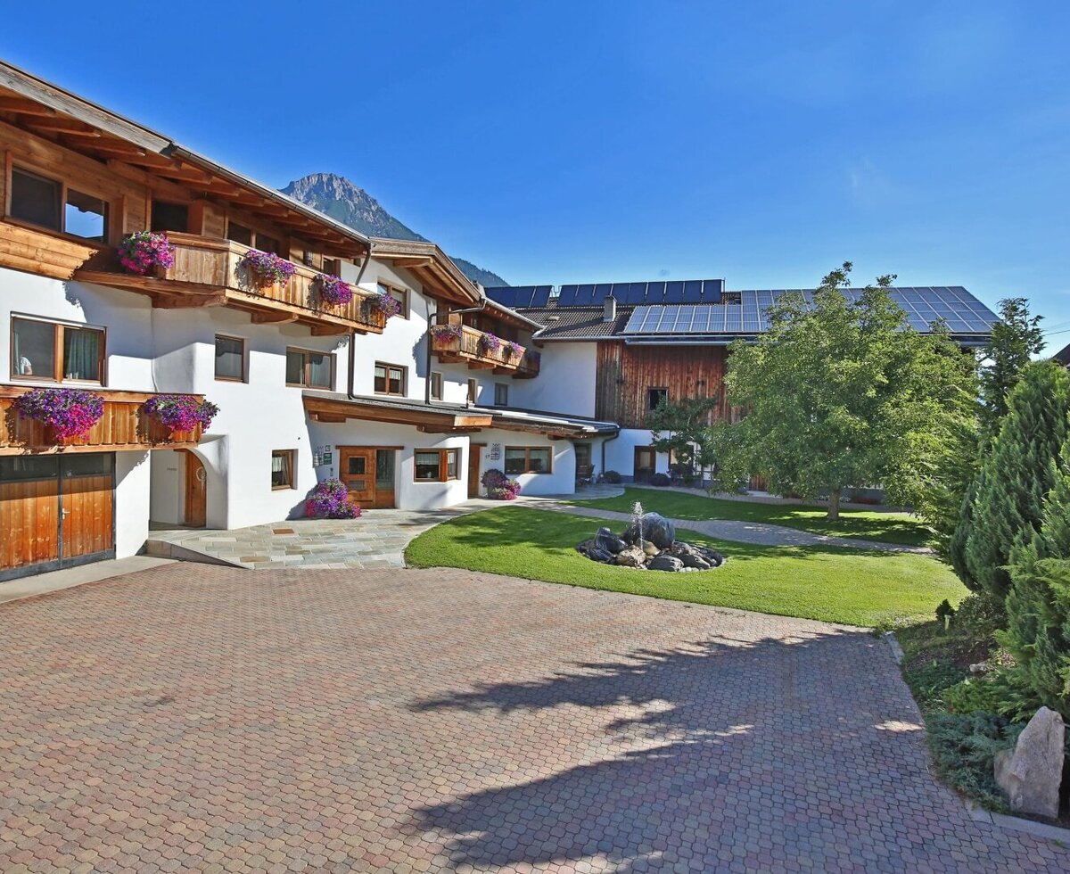 Der Außenbereich des Bauernhofs mit gepflastertem Hof, Garten mit Brunnen, blumengeschmückten Holzbalkonen und Bergblick.