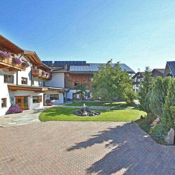Außenansicht des Bauernhofs mit blumengeschmückten Holzbalkonen, gepflastertem Hof und einem Brunnen im Garten.