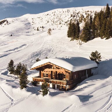 Die Holz-Alm, schneebedeckt und umgeben von Skipisten mit sichtbaren Skispuren und Nadelbäumen.