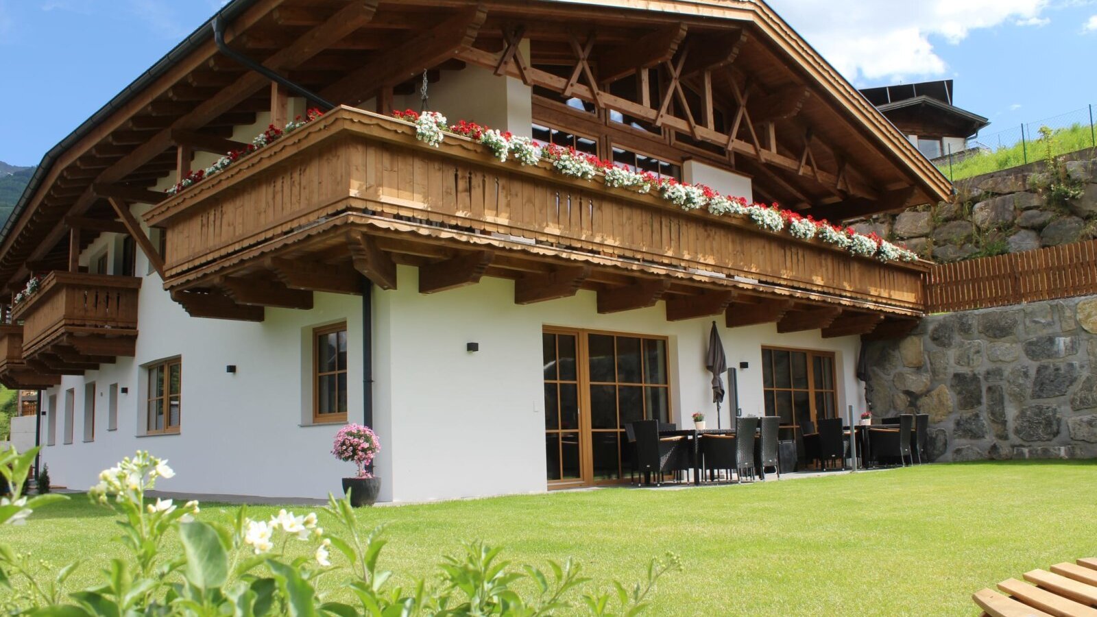 Außenansicht der Ap­par­te­ments mit Holzbalkonen, Blumenkästen, einer Rasenfläche und einem Außenbereich mit Sitzgelegenheiten.