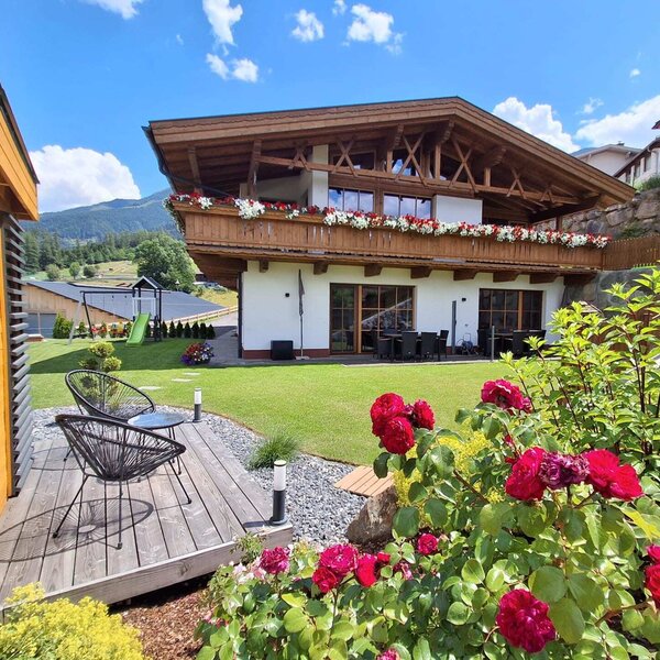 Außenansicht der Ap­par­te­ments mit blumengeschmückten Balkonen, Garten, Außensitzplätzen und Kinderspielplatz.