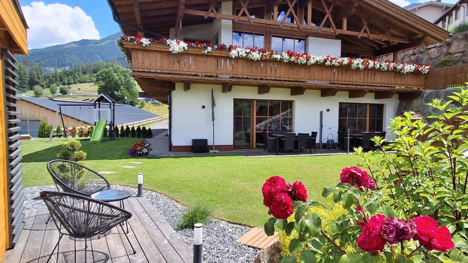Außenansicht der Ap­par­te­ments mit blumengeschmückten Balkonen, Garten, Außensitzplätzen und Kinderspielplatz.