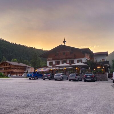 Restaurant neben dem Bauernhof bei Sonnenuntergang, mit Parkplätzen für Gäste und der umliegenden Berglandschaft.