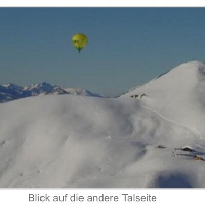Winterlandschaft der Alm mit schneebedeckten Hängen, Skispuren und einem Heißluftballon.