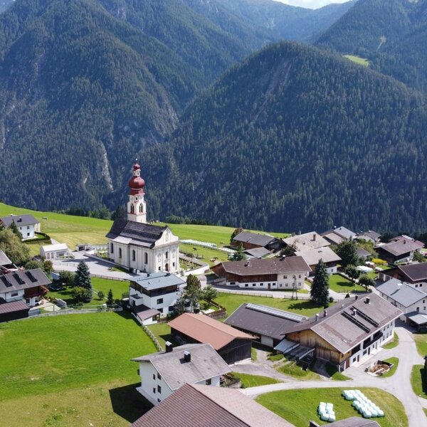 Blick auf das Dorf Asch mit dem Bauernhof, umgeben von grünen Wiesen und Bergen, und einer markanten Kirche.