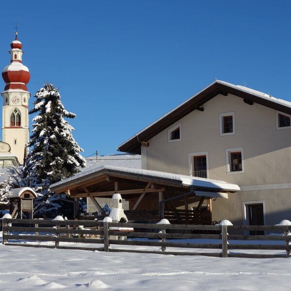 Die Außenansicht des Bauernhofs im Winter, mit schneebedeckter Umgebung, einem Holzzaun und einer Kirche im Hintergrund.
