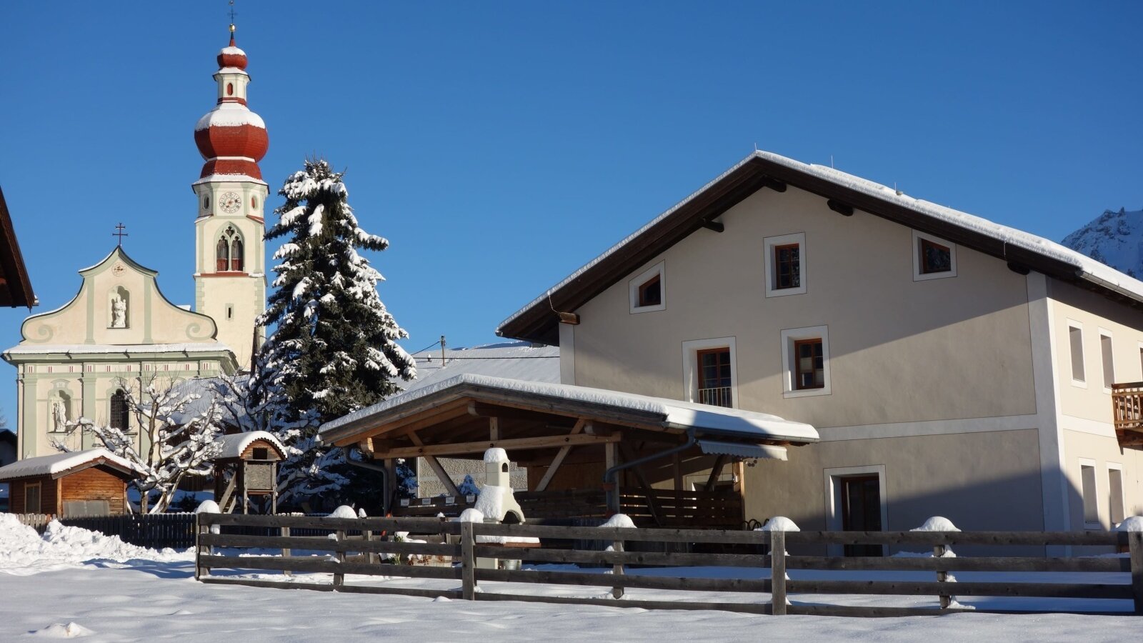 Die Außenansicht des Bauernhofs im Winter, mit schneebedeckter Umgebung, einem Holzzaun und einer Kirche im Hintergrund.