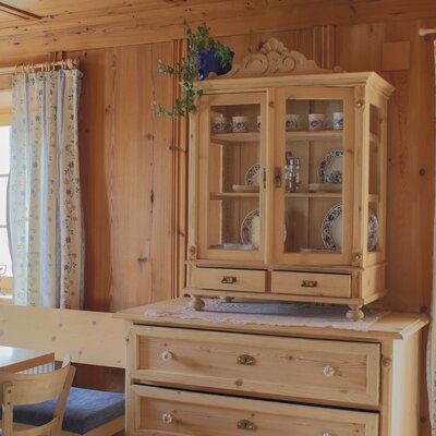 Hölzerner Vitrinenschrank mit Geschirr auf einer Kommode im Essbereich des Bauernhofs, umgeben von Holzverkleidung und Fenstern mit Vorhängen.