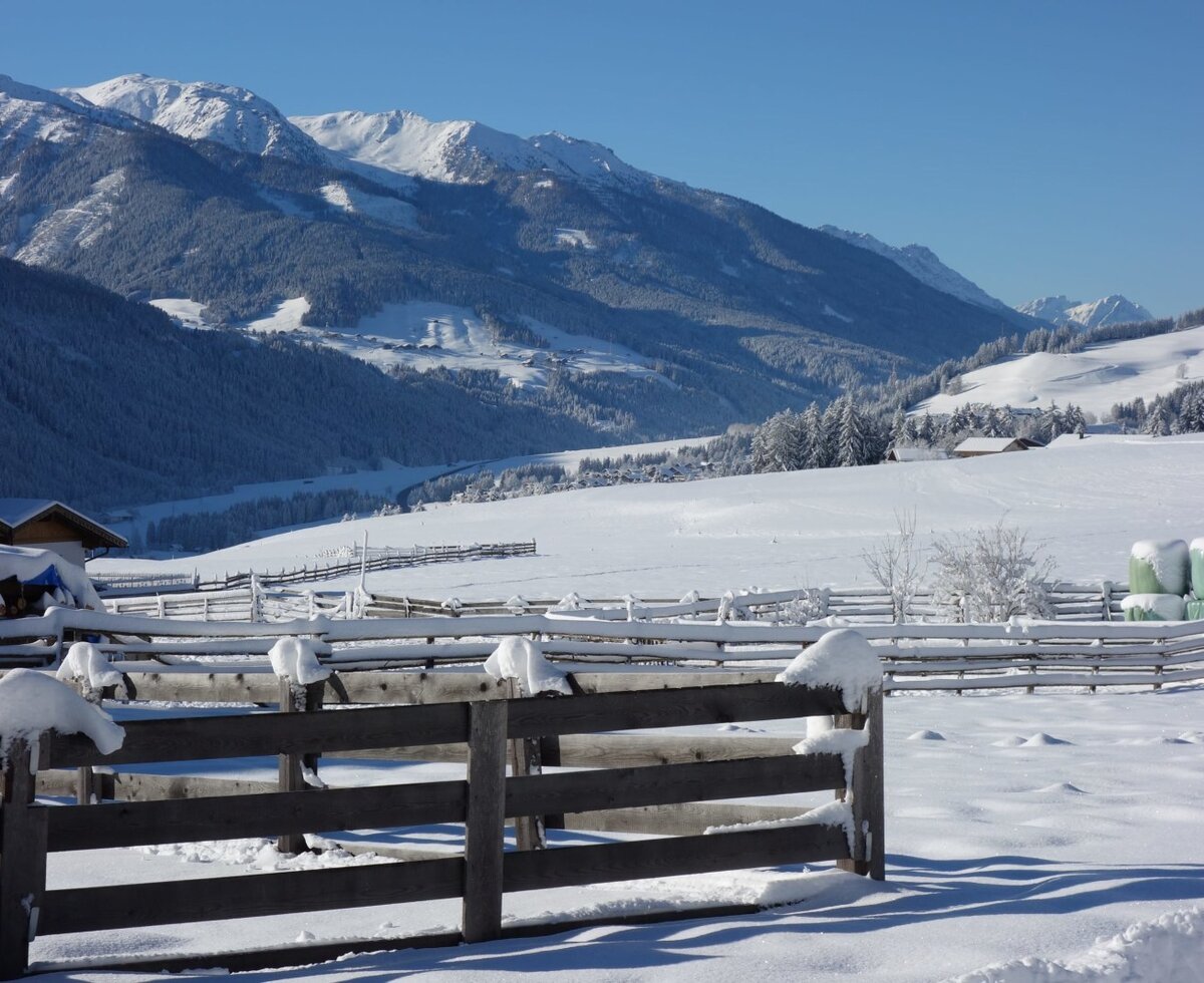 Der verschneite Außenbereich des Bauernhofs mit Holzzäunen, Feldern und Bergen im Hintergrund.
