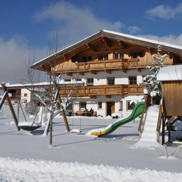 Der winterliche Bauernhof mit einem schneebedeckten Garten, einem Kinderspielplatz mit Schaukel und Rutsche sowie einer Kirche im Hintergrund.