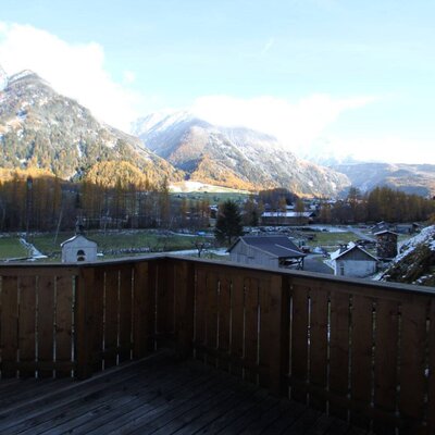 Der Balkon des Bauernhofs mit Holzgeländer bietet einen Ausblick auf das Tal mit schneebedeckten Bergen und herbstlichen Bäumen.