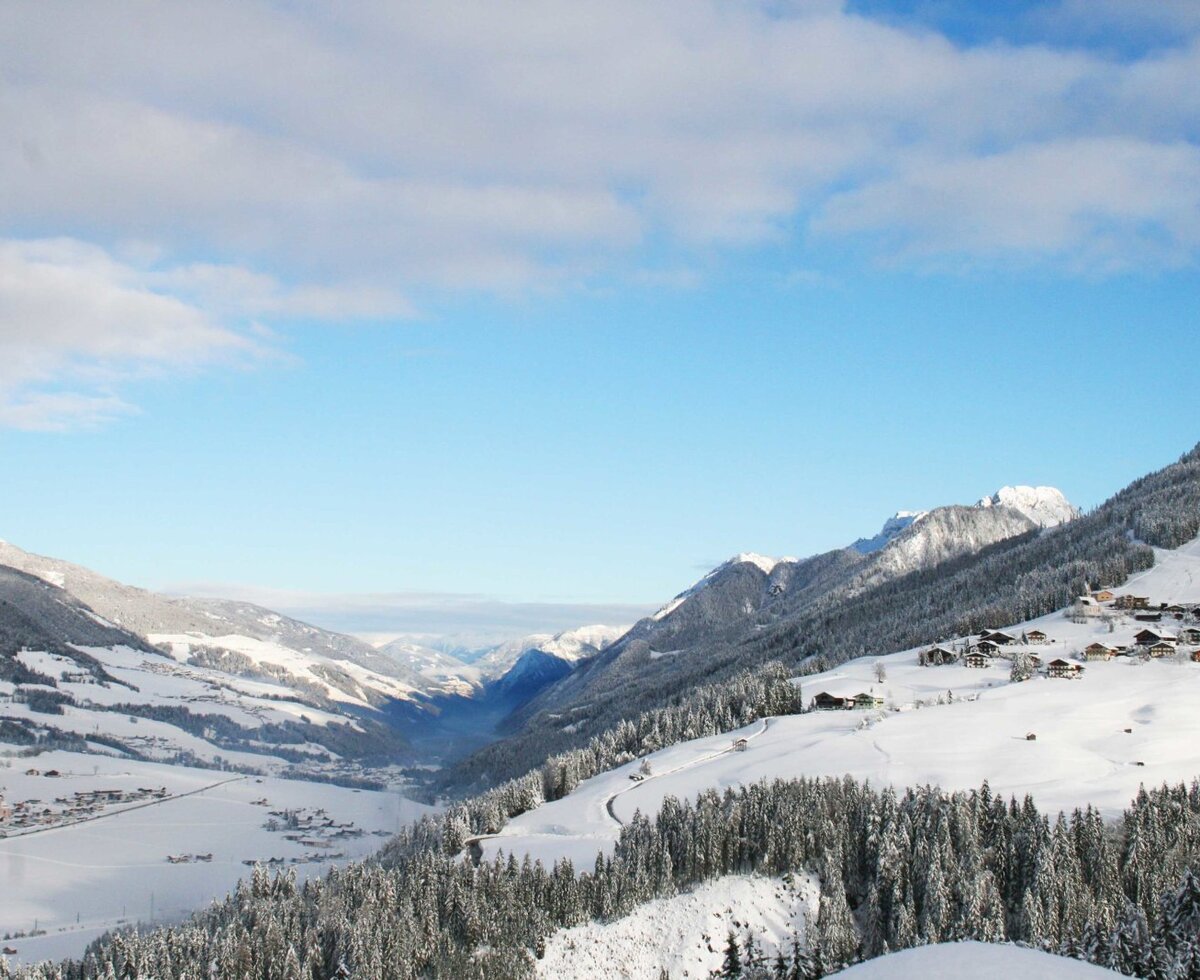 Winterliche Berglandschaft mit verschneitem Tal, einer Siedlung und Skipiste im Umfeld des Bauernhofs.