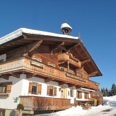 Außenansicht des Bauernhofs mit traditioneller Holzfassade, mehreren Balkonen und einem Glockenturm auf dem Dach, im Winter mit Schnee und Bergblick.