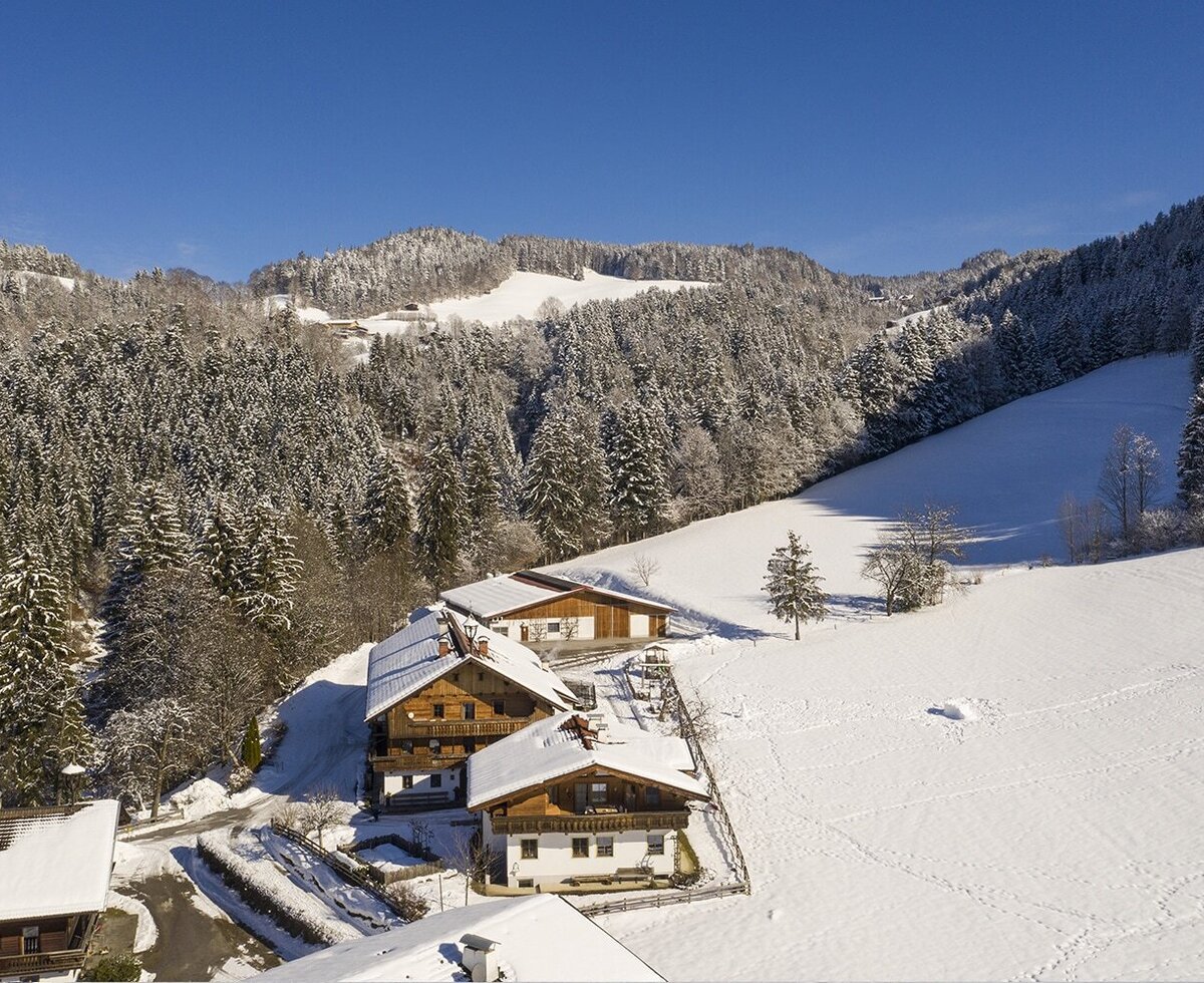 Der Bauernhof in einer schneebedeckten Winterlandschaft, umgeben von verschneiten Wäldern und Bergen.