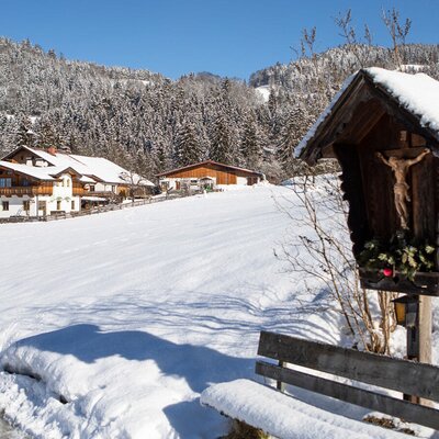 Die Außenansicht des Bauernhofs in einer verschneiten Winterlandschaft, umgeben von Bergen und einem traditionellen Wegkreuz.