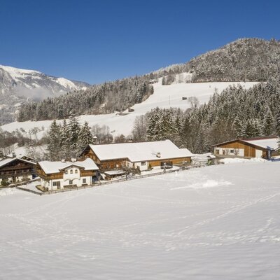 Die Außenansicht des Bauernhofs im Winter, umgeben von verschneiten Feldern, Wäldern und Bergen.