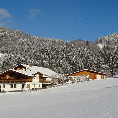 Der Bauernhof und eine Scheune in einer verschneiten Winterlandschaft unter blauem Himmel, umgeben von Wäldern.
