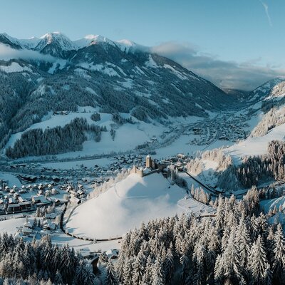 Blick auf das verschneite Tal mit Dorf und Burg, umgeben von schneebedeckten Bergen.