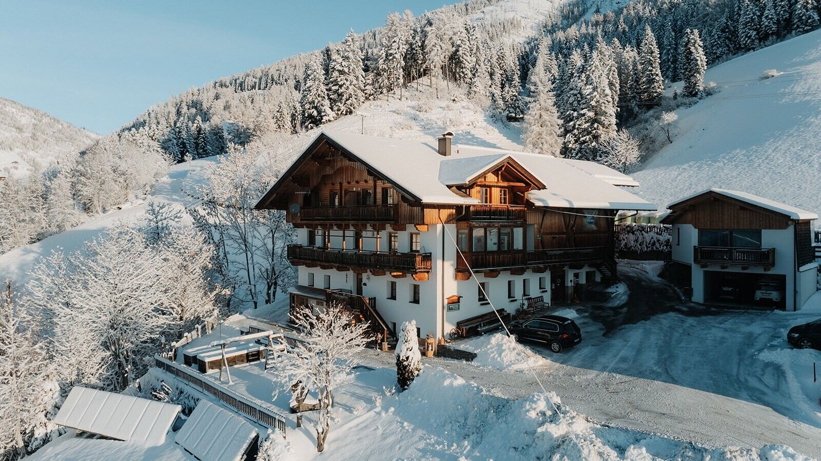 Der Bauernhof im Winter mit Balkonen und einem Kinderspielplatz auf der Terrasse, umgeben von verschneiten Bergen.
